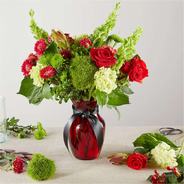 Red roses and green flowers arranged in a red glass vase with a black ribbon.