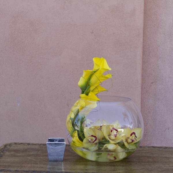 Yellow orchids arranged in a round glass bowl with a small square candle holder beside it