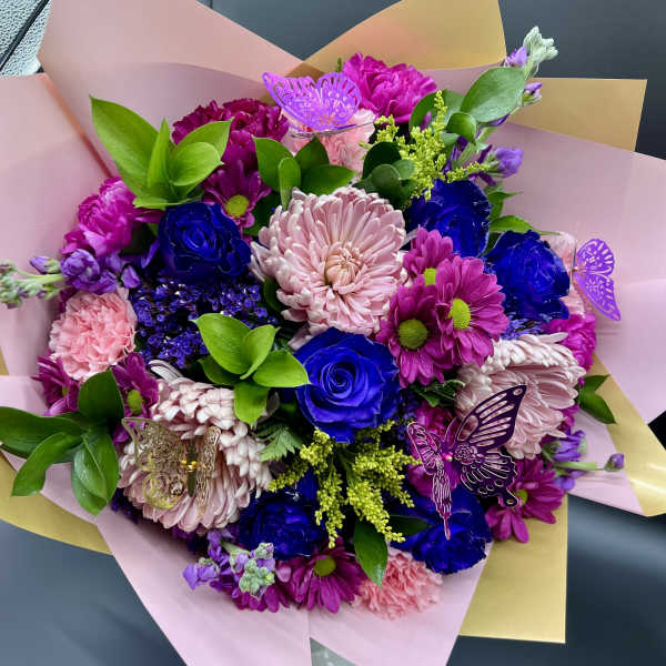 Hand-tied bouquet of pink mums and blue roses wrapped in pink and yellow paper with butterfly picks