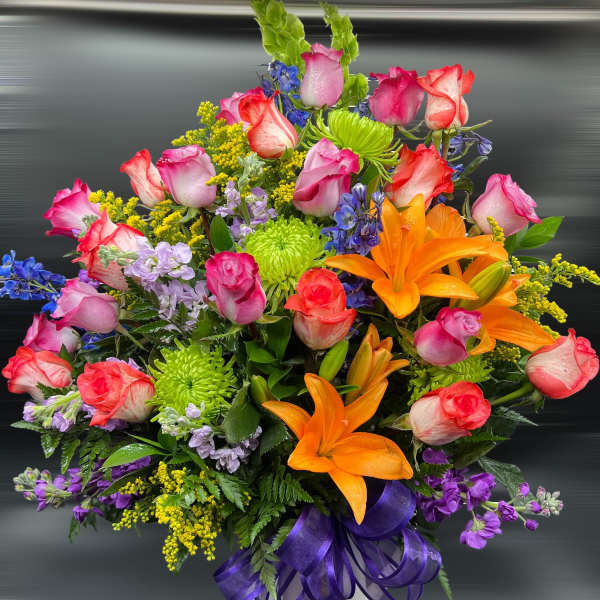 Tall mixed arrangement of pink roses, orange lilies, green mums, and blue accents in a white vase with purple ribbon.