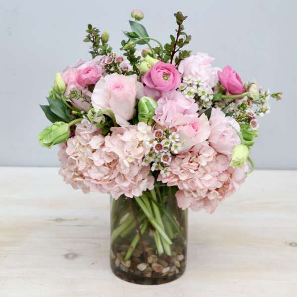 Round arrangement of pink hydrangeas, roses, and ranunculus in a clear glass vase with pebbles.