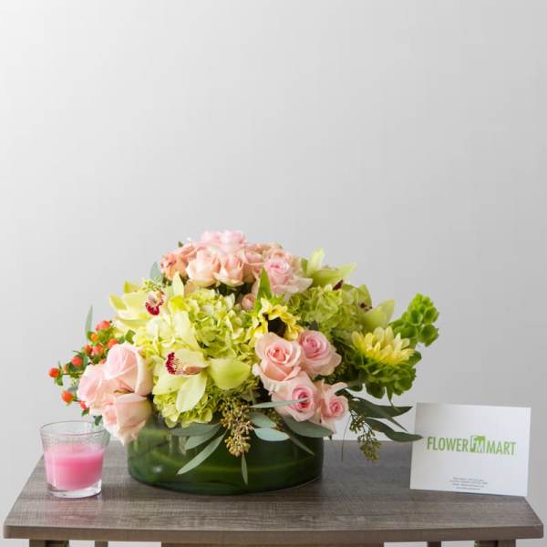 Pink and green floral arrangement in a low glass vase with a candle and card