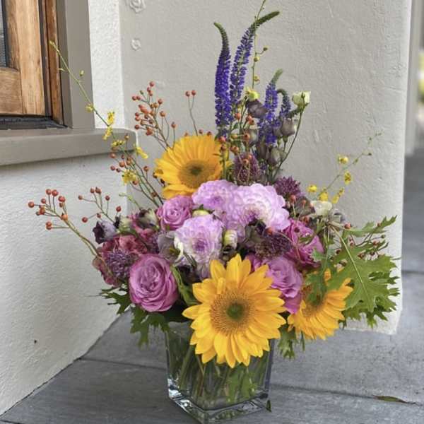 Mixed bouquet in a clear glass vase with sunflowers, purple roses, and tall blue spikes