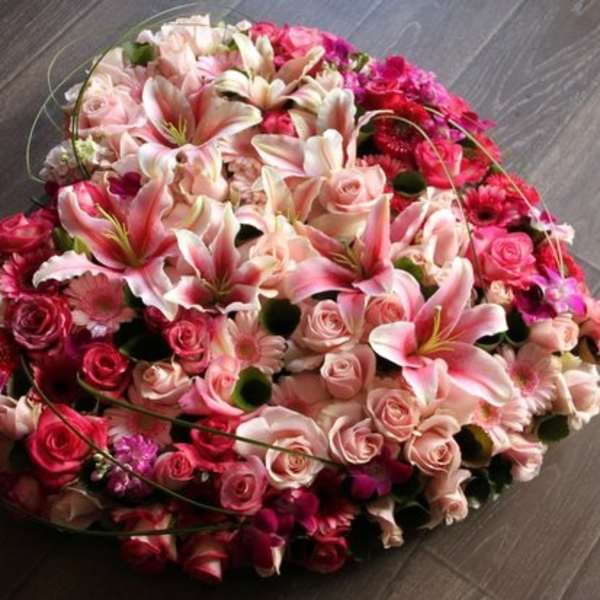 Heart-shaped arrangement of pink lilies, roses, and gerbera daisies in various pink and red tones