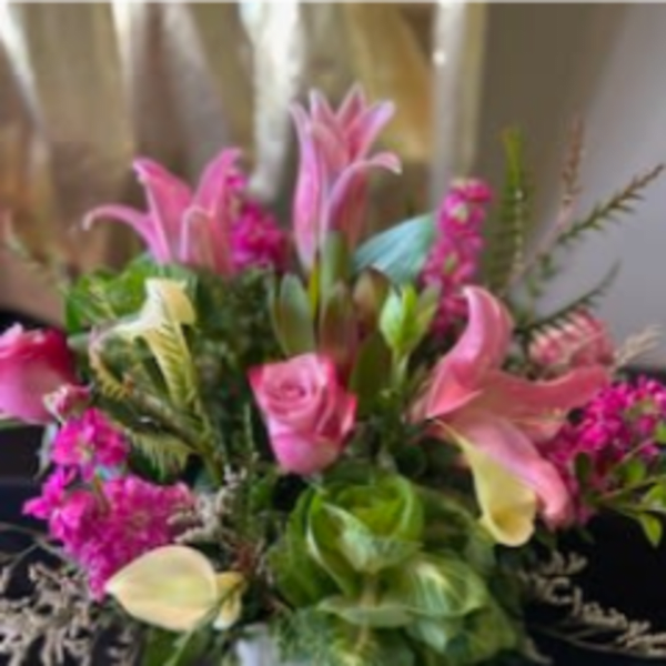 Pink lilies, roses, and pale calla lilies arranged with bright filler flowers in a white vase