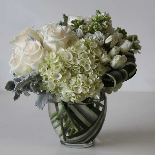 Compact arrangement of white roses and hydrangeas in a clear glass vase with looped striped leaves