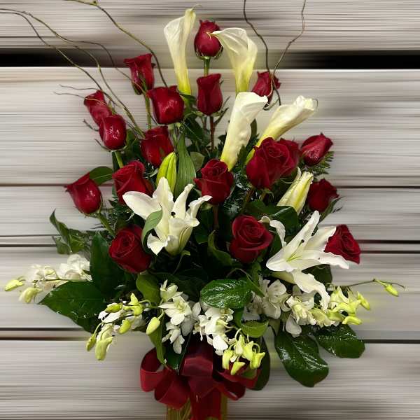Tall arrangement of red roses, white lilies, and calla lilies in a glass vase with a red ribbon