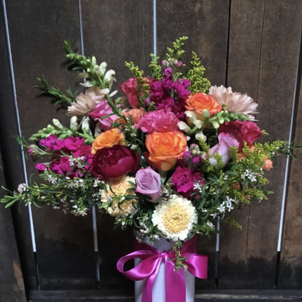 Mixed bouquet of pink, orange, and white flowers in a tall white vase with a pink ribbon