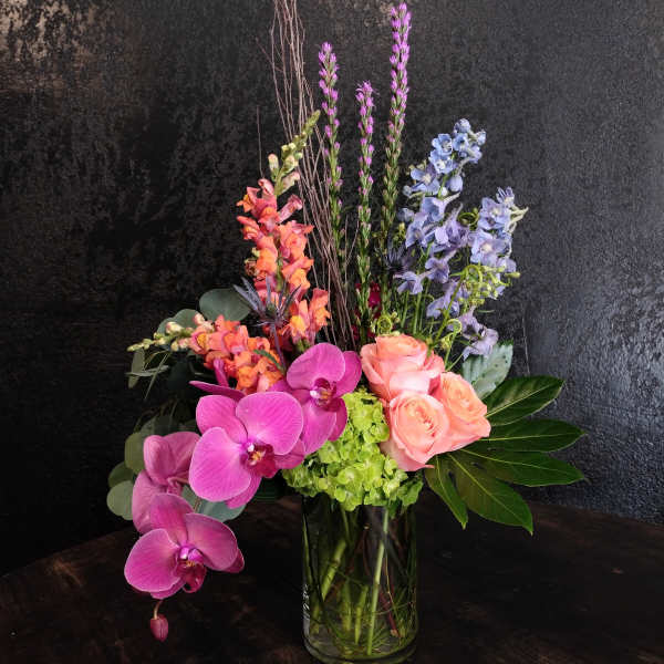 Tall modern arrangement with pink orchids, peach roses, blue delphinium and green hydrangea in a glass vase