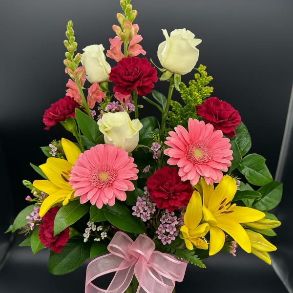 Mixed bouquet with pink gerbera daisies, yellow lilies, white roses, and red carnations in a vase with a pink bow.