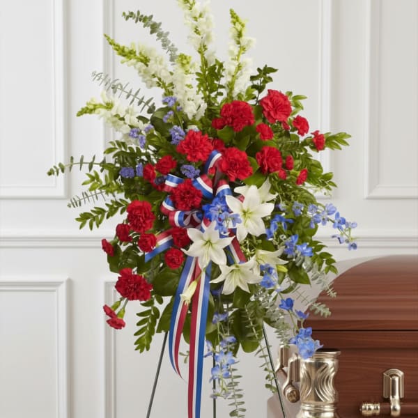 Tall red, white, and blue standing spray with carnations, lilies, and ribbon on an easel beside a casket.