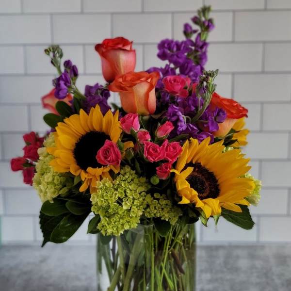 Bright mixed arrangement with sunflowers, roses, and purple blooms in a clear glass vase