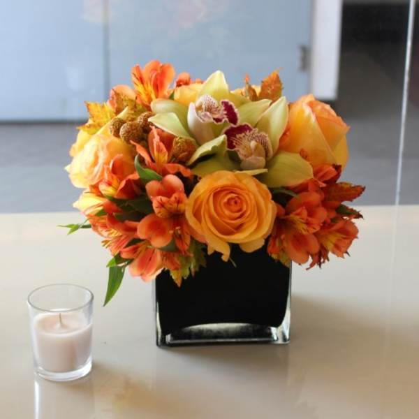 Orange and yellow floral arrangement in a black square vase beside a small candle.