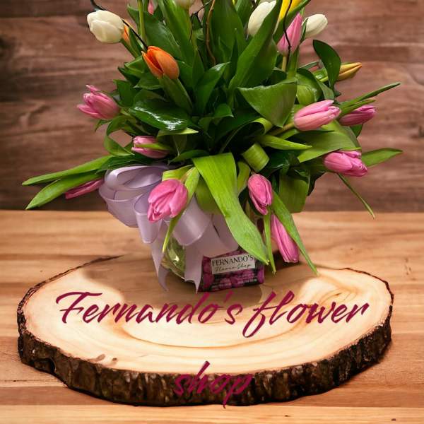 Mixed tulip arrangement with colorful butterfly picks in a clear vase on a wood slice base.