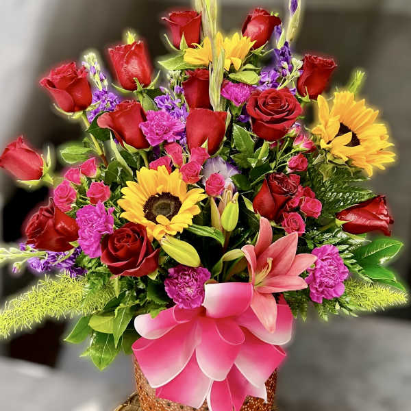 Tall mixed arrangement with red roses, sunflowers, pink lilies, and carnations in an orange pot with a pink bow.