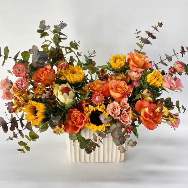 Mixed bouquet of orange roses, sunflowers, and pink blooms in a white vase