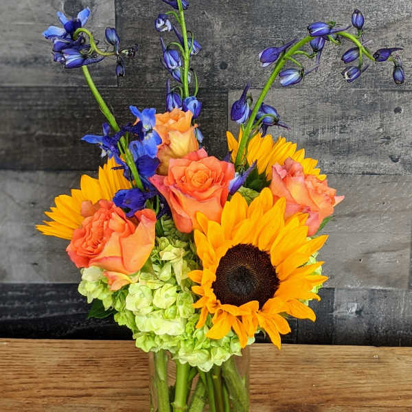 Tall glass vase with yellow sunflowers, orange roses, green hydrangeas, and blue delphinium.