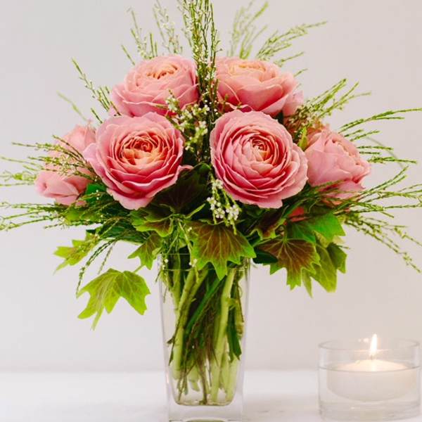 Pink roses arranged in a clear glass vase with greenery and a lit candle nearby