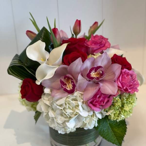 Compact arrangement of white hydrangeas, red roses, pink orchids, tulips, and calla lilies in a glass vase.
