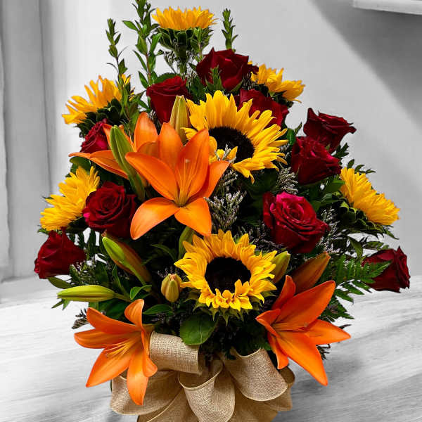 Mixed arrangement of sunflowers, orange lilies, and red roses in a glass vase with a burlap bow