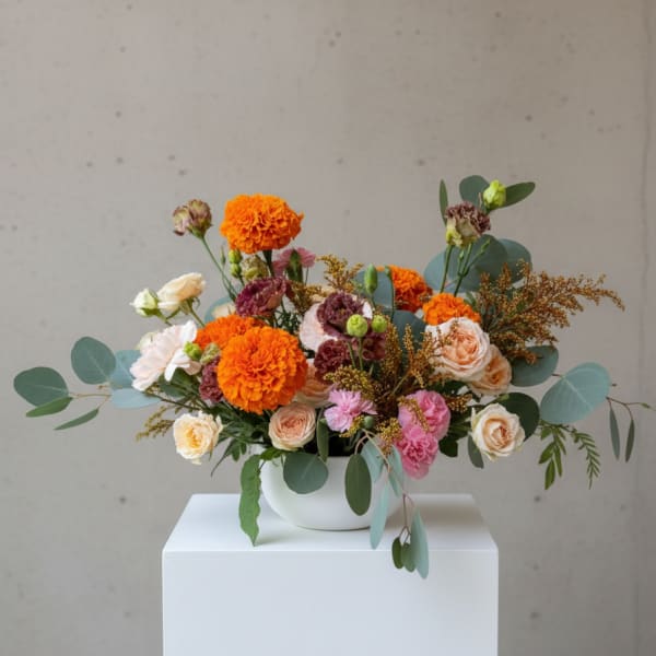 Low arrangement of orange marigolds and blush roses in a white bowl vase on a white pedestal.