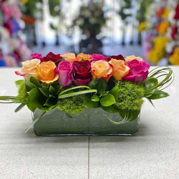 Low rectangular arrangement of mixed pink, orange, yellow, and red roses with green moss and looped grass in a glass vase.