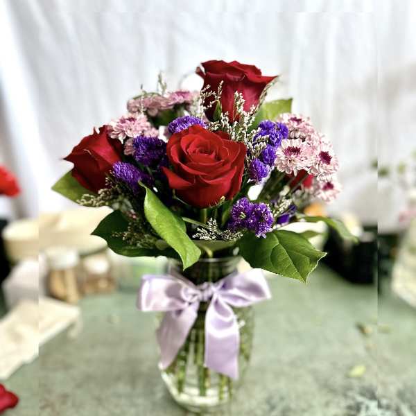 Red roses and purple filler flowers in a glass vase with a lavender ribbon