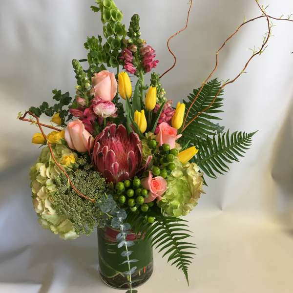 Tall mixed bouquet in a glass vase with pink roses, yellow tulips, and a protea