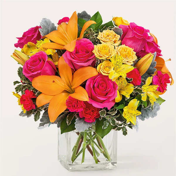 Bright mixed bouquet of orange lilies, pink roses, and yellow blooms in a clear glass cube vase