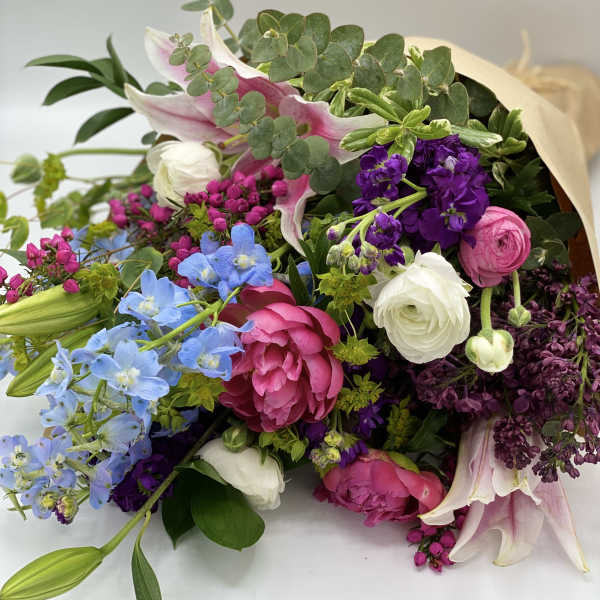 Colorful hand-tied bouquet with pink peonies, blue delphinium, white blooms and lilies wrapped in kraft paper.