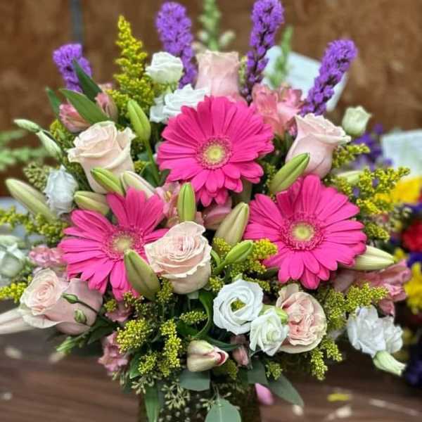Vase arrangement with hot pink gerbera daisies, pale roses, lilies, and mixed blooms in a green textured glass vase