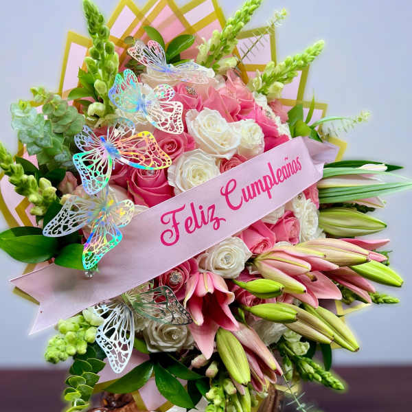 Tall arrangement of pink lilies and pink and white roses with a Feliz Cumpleaños ribbon and butterfly accents.