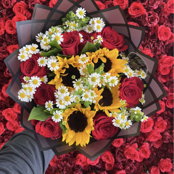 Handheld bouquet of red roses, yellow sunflowers, and white daisies wrapped in black paper over a bed of red roses.