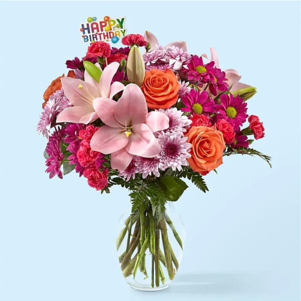 Mixed bouquet of pink lilies, orange roses, and purple mums in a clear glass vase with a Happy Birthday pick