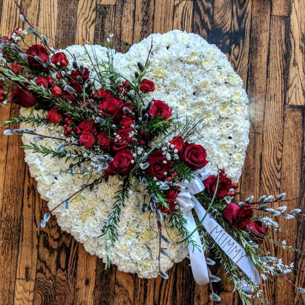 Heart-shaped white funeral arrangement with red rose spray and 'MUMMY' ribbon on a wooden floor