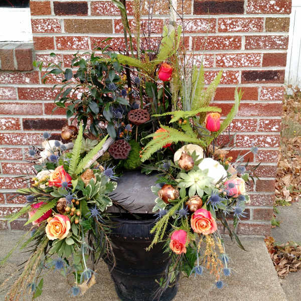 Outdoor arrangement of orange roses, succulents, and pods in a black urn against a brick wall.