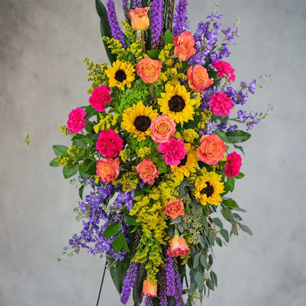 Tall standing spray of sunflowers, orange roses, pink carnations, and purple blooms on an easel.