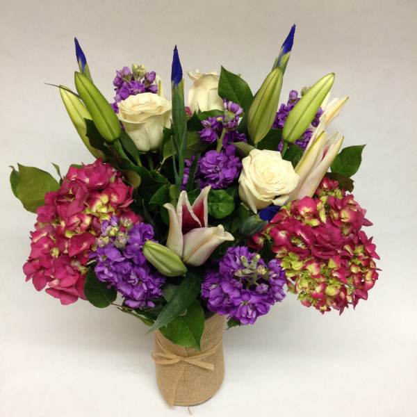 Bouquet of white roses, purple blooms, and lily buds in a burlap-wrapped vase
