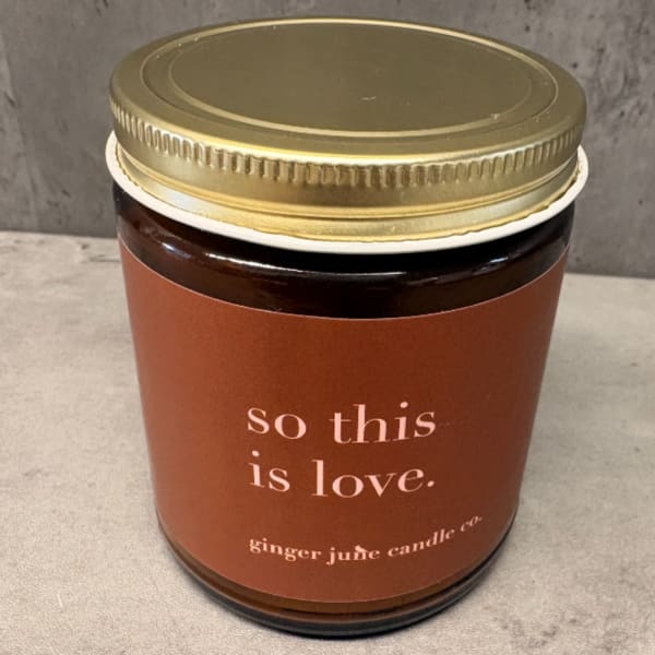 Amber glass jar candle with gold lid and brown label reading 'so this is love.'