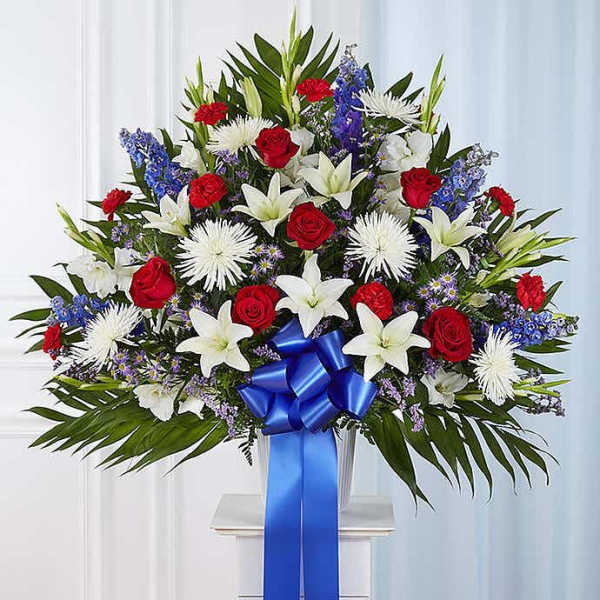 Tall red, white, and blue sympathy arrangement with roses, lilies, mums, and a large blue ribbon bow