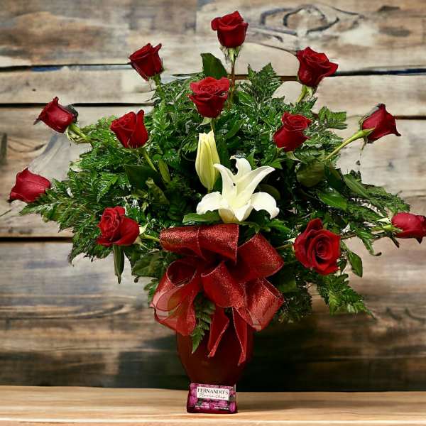 Tall arrangement of red roses and white lilies in a red vase with red bow and chocolates