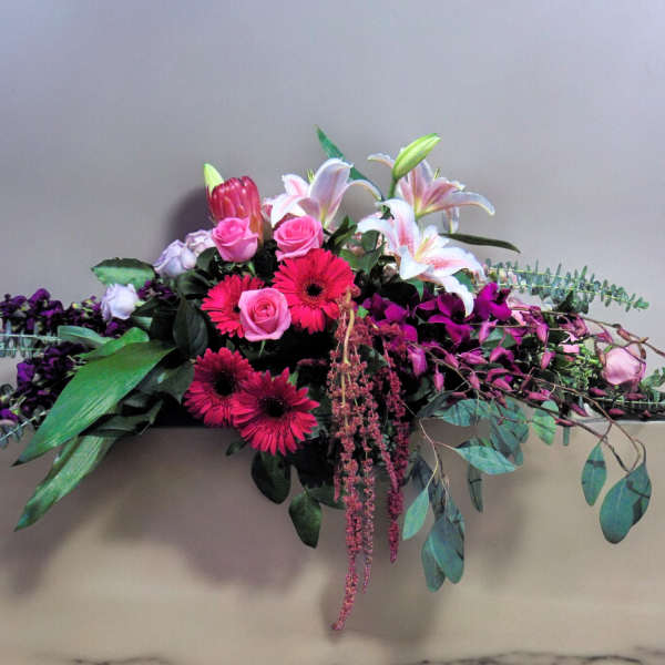Long low arrangement with pink gerbera daisies, roses, lilies and purple blooms