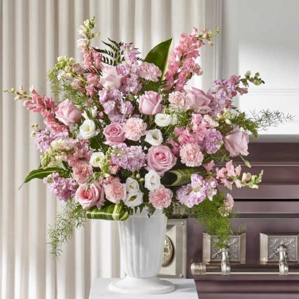 Tall pink and white arrangement with roses and carnations in a white pedestal vase beside a casket.
