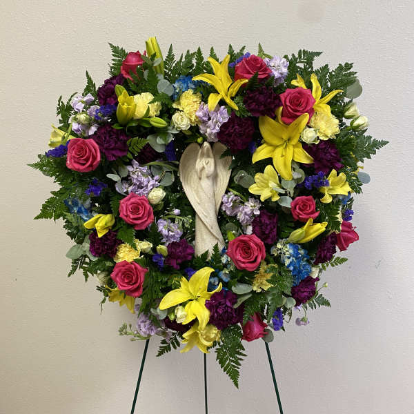 Heart-shaped floral wreath with roses, lilies, and a central angel figure on a stand