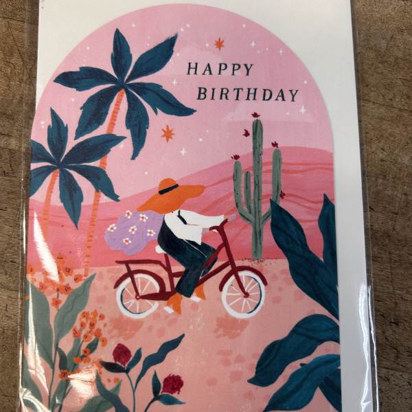 Illustrated Happy Birthday card with a person riding a bicycle through a pink desert scene