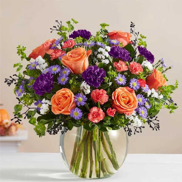 Mixed bouquet of orange roses, purple carnations, and small daisies in a round clear glass vase