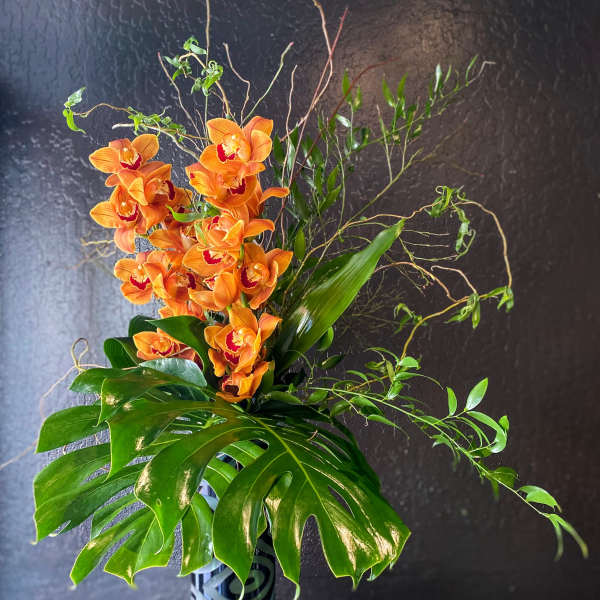 Tall arrangement of orange orchids with large monstera leaves in a patterned black cylinder vase