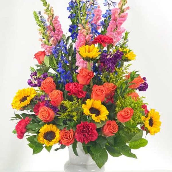 Tall mixed arrangement of sunflowers, orange roses, pink snapdragons and purple flowers in a white vase