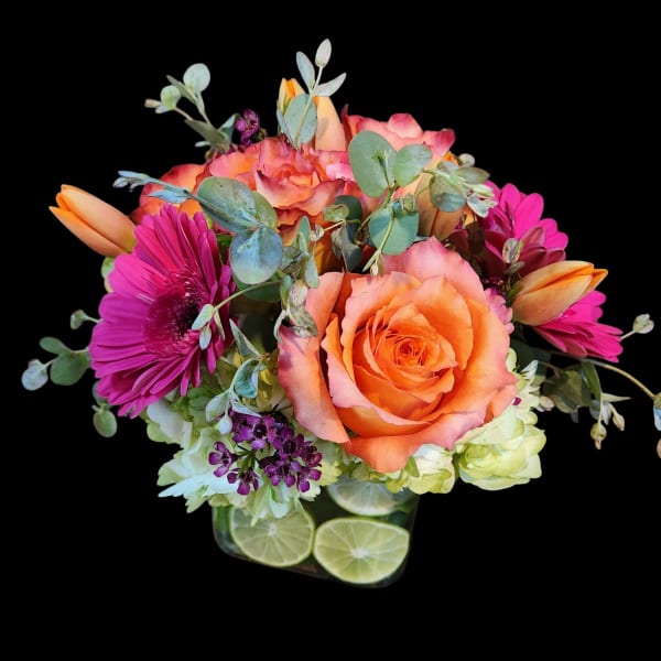 Bright arrangement of orange roses, hot pink gerbera daisies and tulips in a cube vase lined with lime slices