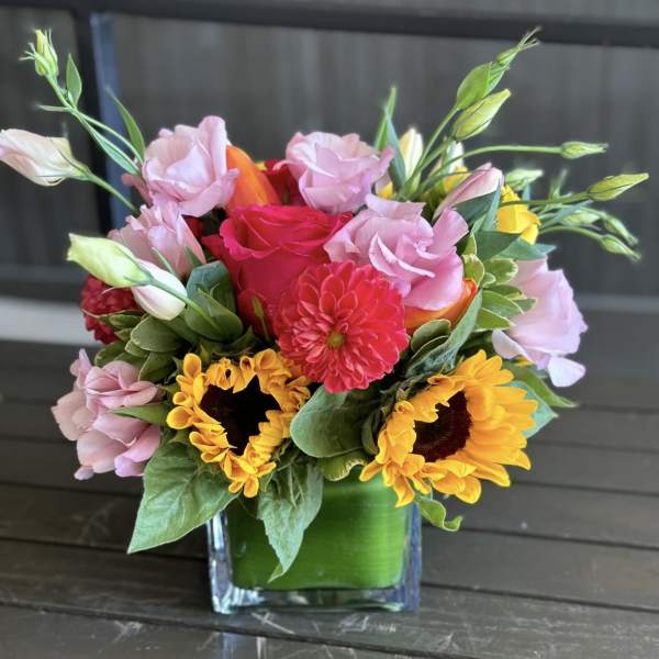 Mixed bouquet of pink, yellow, and red flowers in a square glass vase